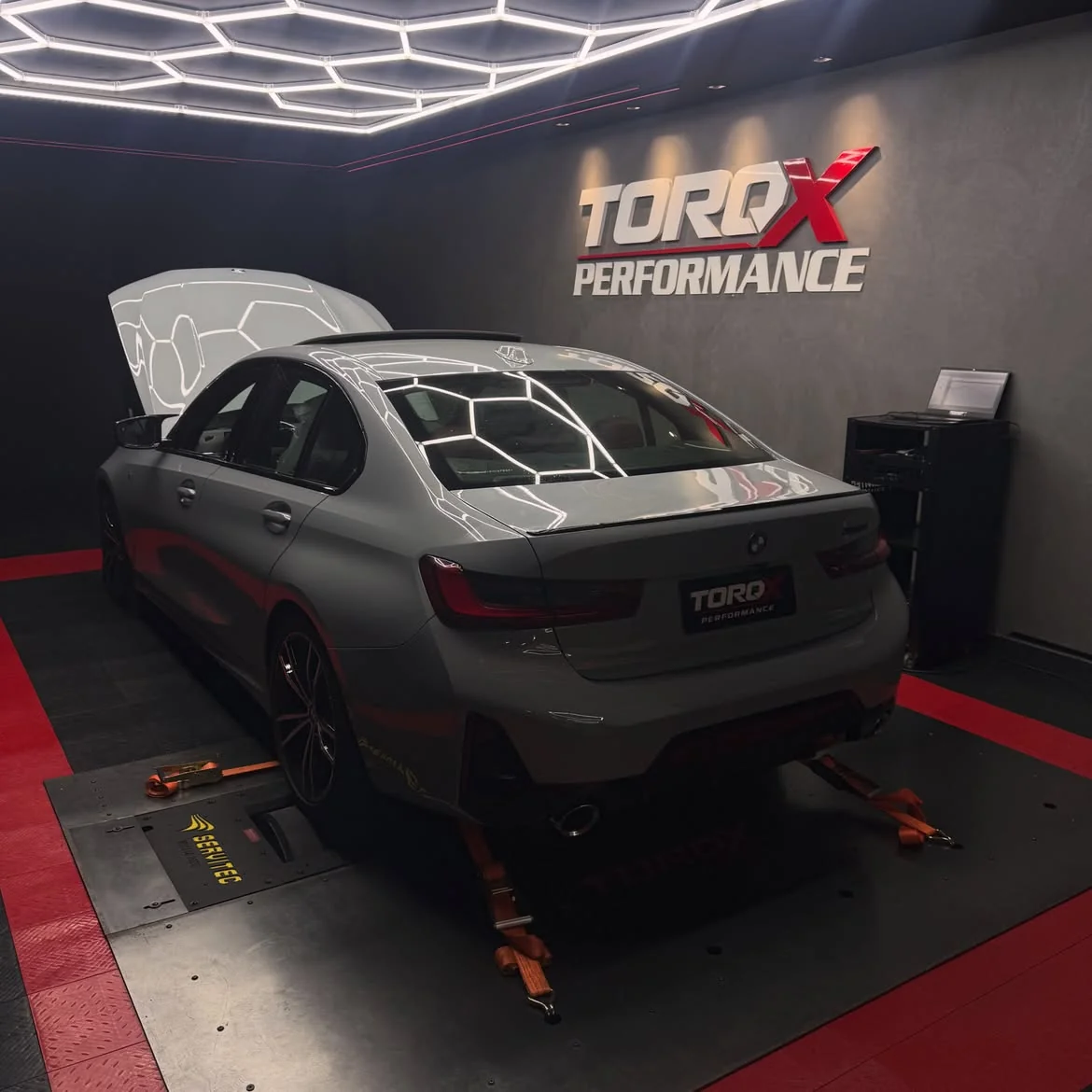 BMW 320i G20 cinza em três quartos traseiro no dinamômetro, com o capô levantado e o letreiro TORQX ao fundo