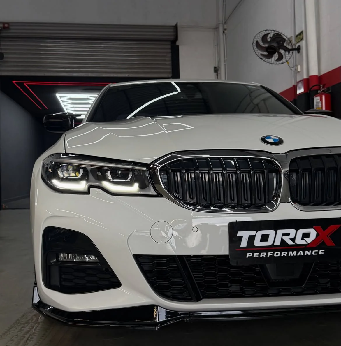 Frente de um BMW 320i G20 branco com front lip e grade dupla escurecida, dentro da oficina