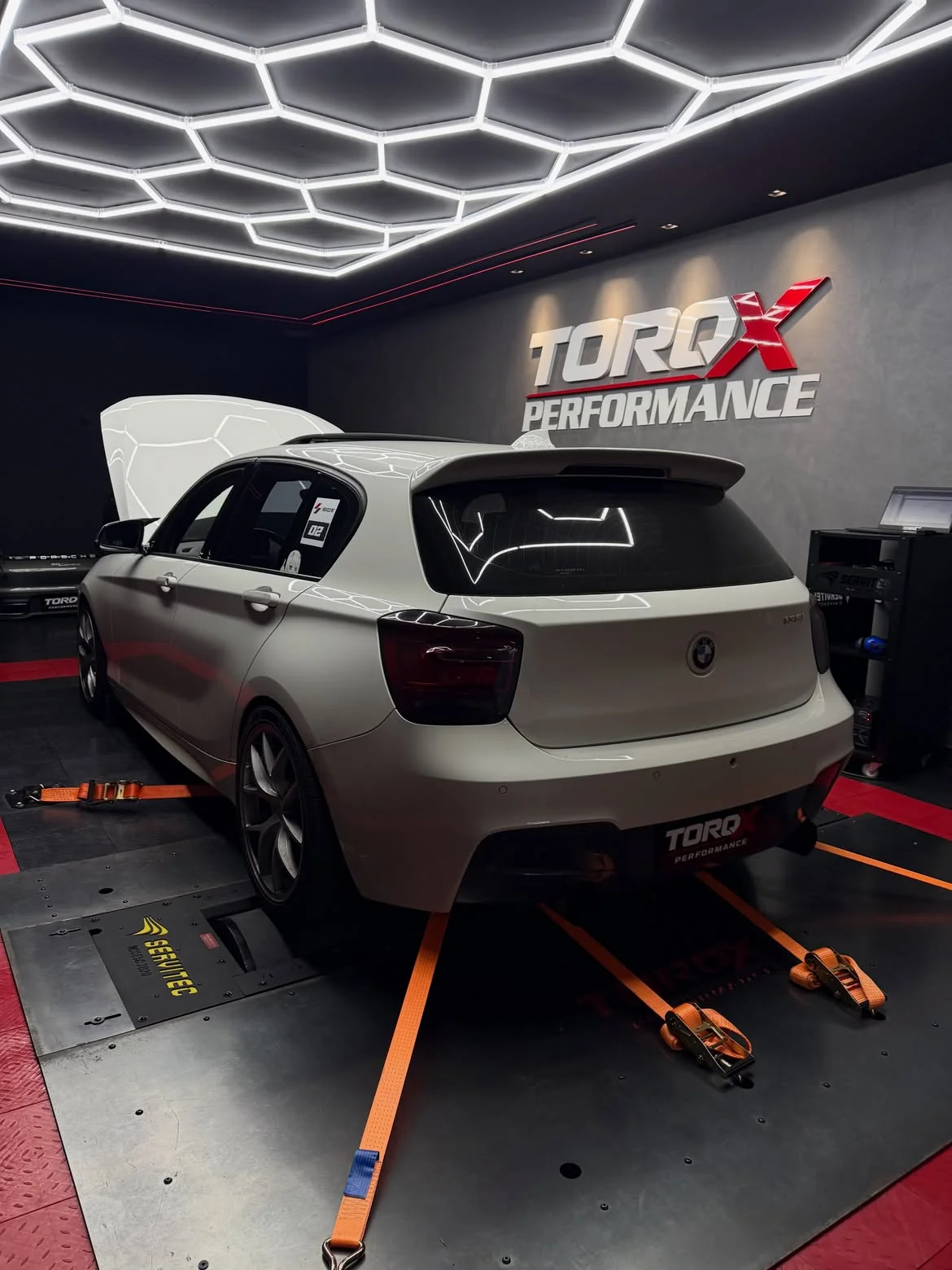 BMW M135i branco em três quartos traseiro no dinamômetro, com cintas laranja cruzadas sob a carroceria