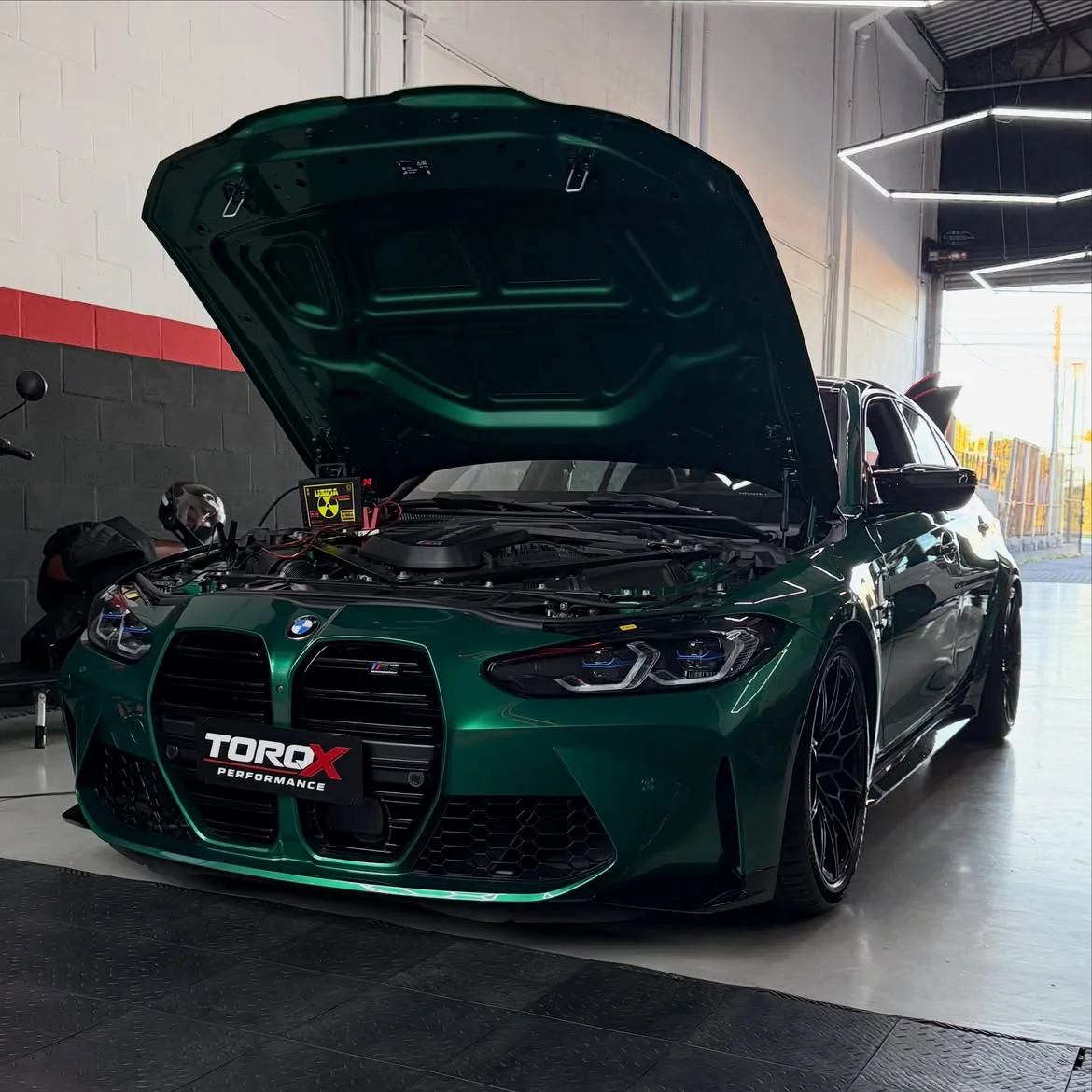 BMW M3 G80 verde-britânico com o capô levantado e mantenedor de bateria ligado ao vão do motor durante a sessão de coding