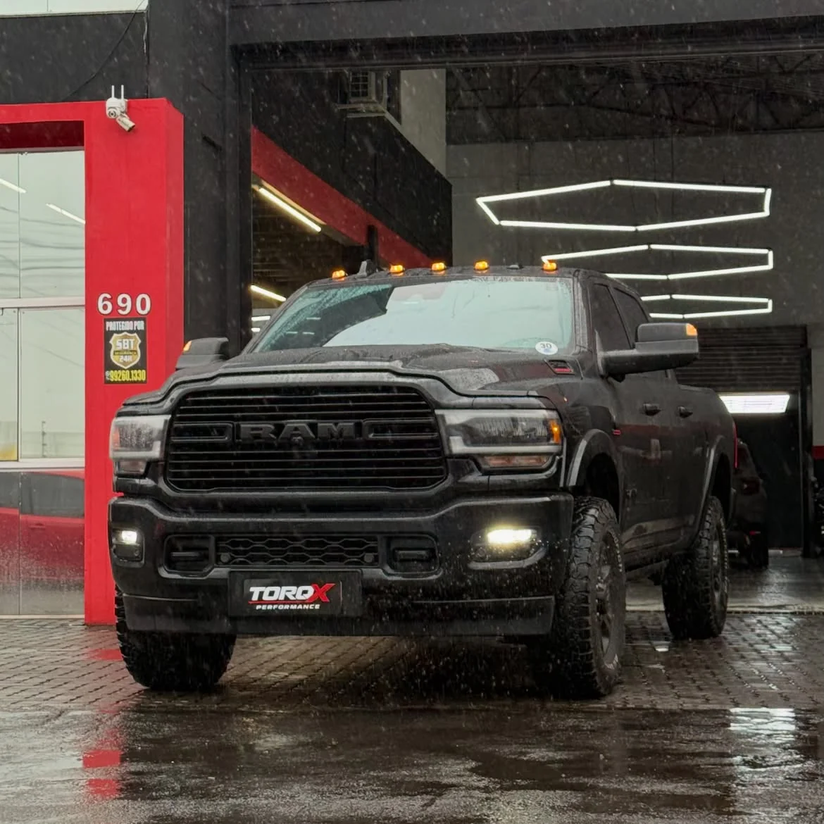 RAM 2500 preta sob chuva na entrada da TORQX, com faróis e luzes de teto acesos