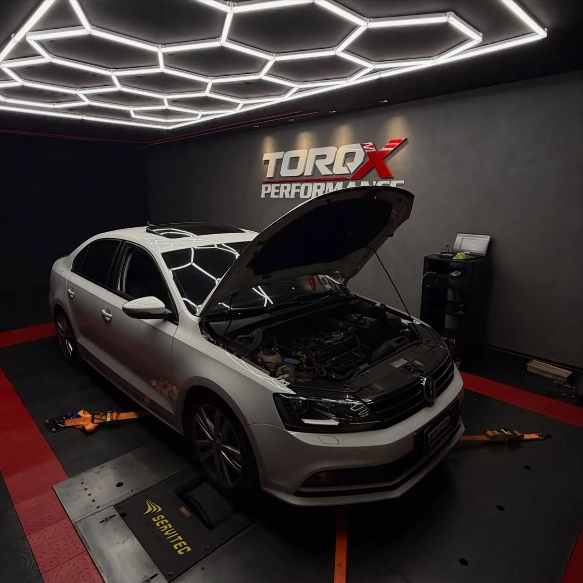 Volkswagen Jetta branco com o capô levantado sobre o leito do dinamômetro, sob a luminária hexagonal