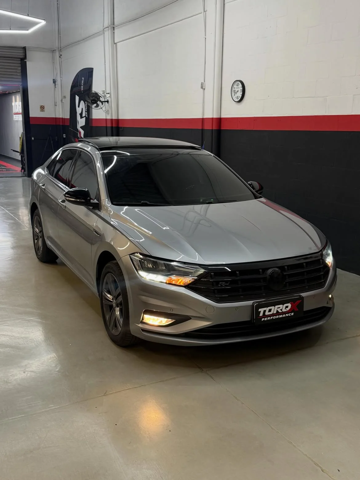 Volkswagen Jetta R-Line prata de três quartos dianteiro com os faróis acesos, dentro da oficina