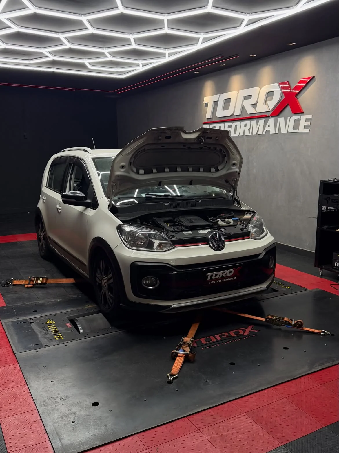 Volkswagen Up branco com o capô levantado sobre o leito do dinamômetro, sob a luminária hexagonal