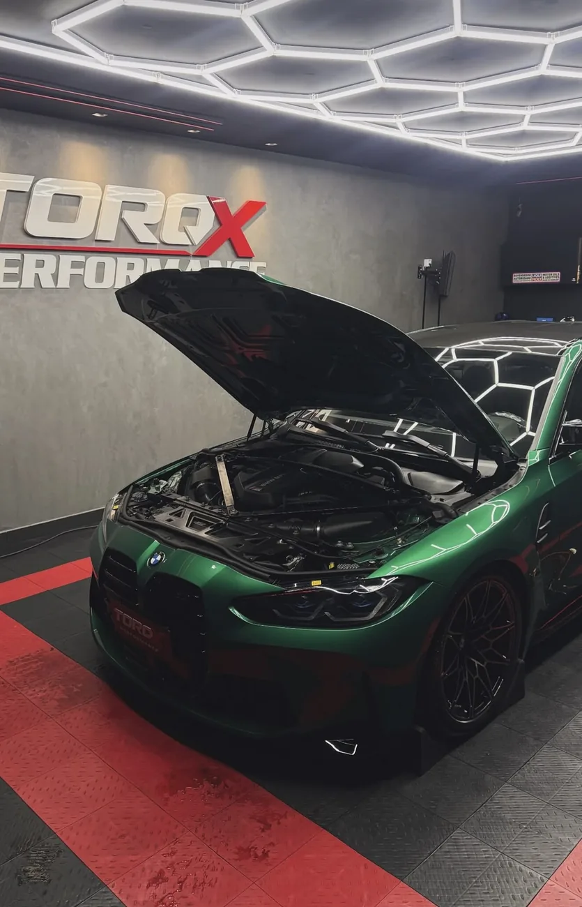 BMW M4 verde com o capô levantado na sala da TORQX, sob a luminária hexagonal e o letreiro da parede